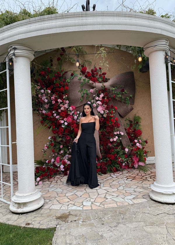 Compleu Bling Miray negru elegant, set 3 piese cu pantaloni evazați și mantou, purtat de femeie în fața unui decor cu flori roșii