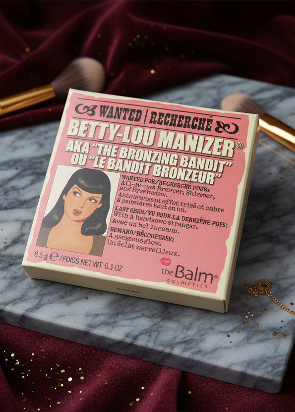 Iluminator și bronzer multifuncțional The Balm Betty-Lou Manizer pe masă de machiaj, ambalaj dreptunghiular roz